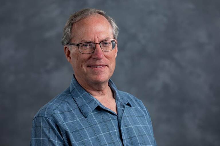 A man with glasses and short gray hair is wearing a blue plaid shirt. He is smiling slightly and positioned against a gray mottled background.