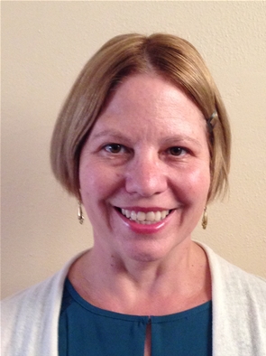 A woman with short blonde hair and a hair clip smiles at the camera. She is wearing a blue top and a light cardigan against a plain background.