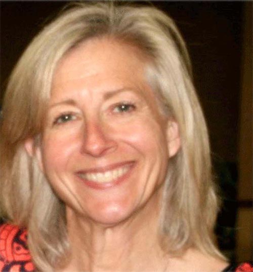 A woman with shoulder-length blonde hair smiling at the camera. She is wearing a red and black patterned top. The background is blurred.