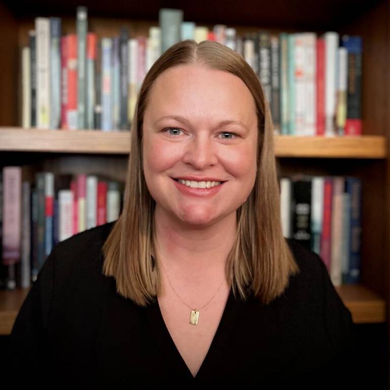 A person with shoulder-length hair smiles in front of a bookshelf filled with books. They are wearing a black top and a gold pendant necklace.