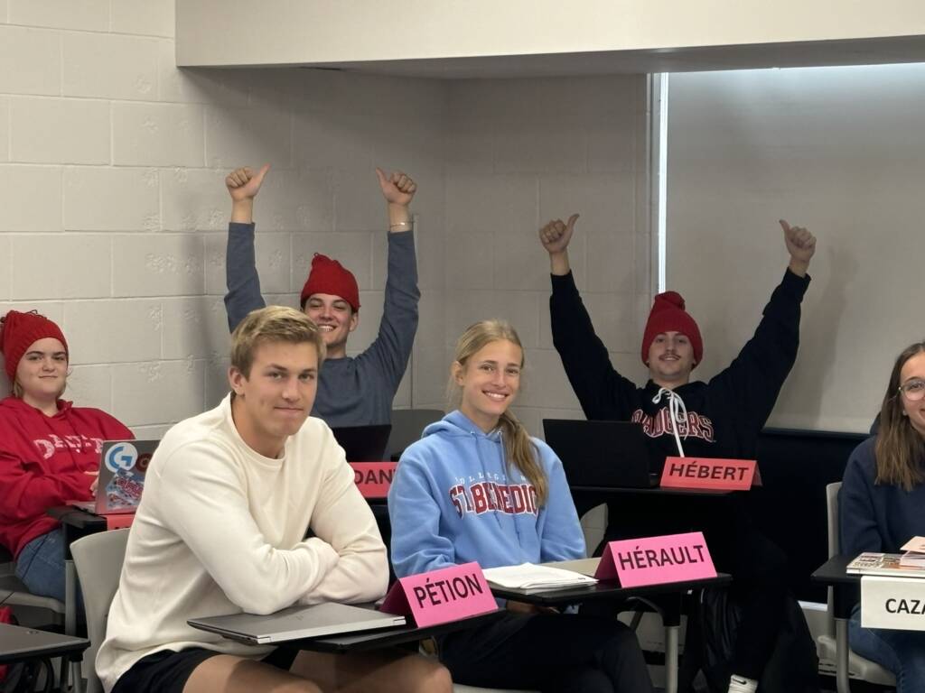 A group of students in a classroom, some wearing red hats. They sit at desks with nameplates, smiling and raising their hands in excitement. Laptops are open on the desks, and they appear engaged in the learning environment.