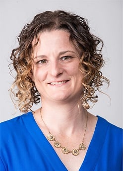 A person with curly hair smiles, wearing a blue top and a decorative necklace, posing in front of a plain background.