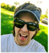 A person wearing sunglasses and a visor is making a playful face in a sunny outdoor setting with green grass in the background.