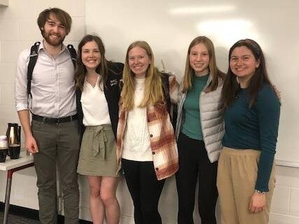 Five people stand smiling in front of a whiteboard. There is a table with coffee supplies to their left. They are casually dressed in a mix of pants, shorts, and skirts. The room has a warm, friendly atmosphere.