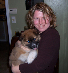 A person with wavy hair is smiling and holding a fluffy brown and white puppy. They are indoors, standing in front of a green wall with a thermostat.