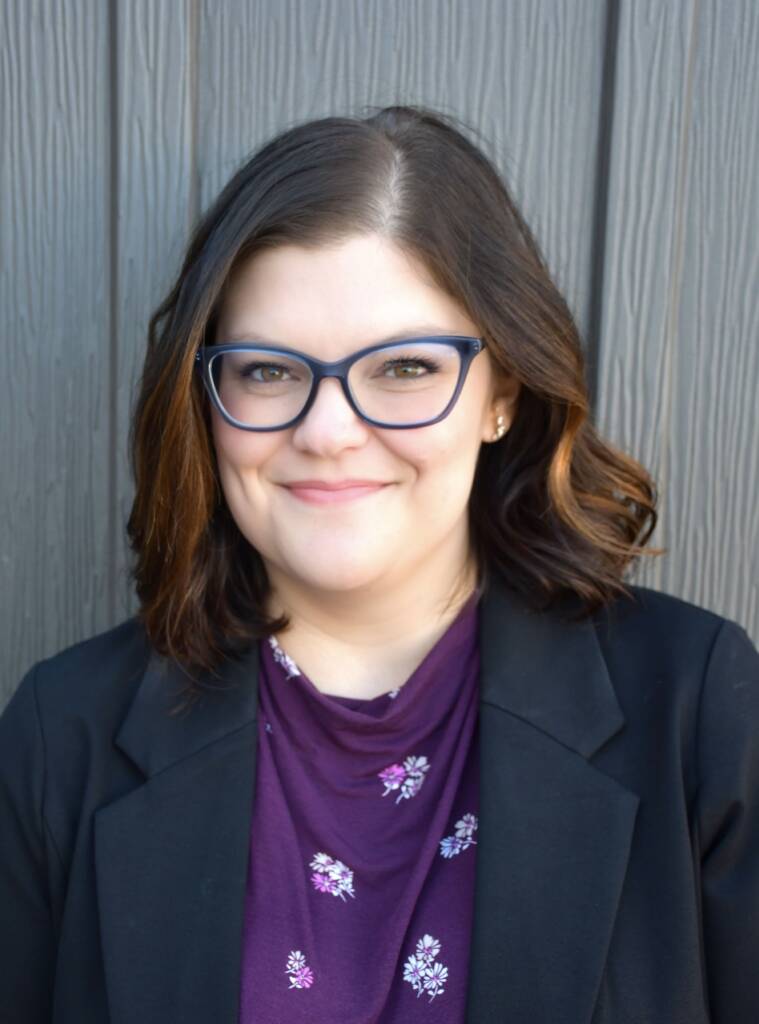 A person with shoulder-length brown hair and glasses smiles at the camera, wearing a black jacket over a purple top with floral designs, standing against a wooden background.