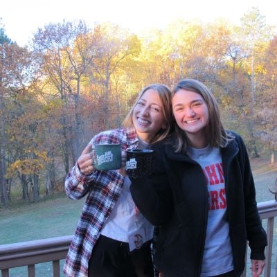 Two female college students standing on a deck with mugs smiling with fall trees in the distance