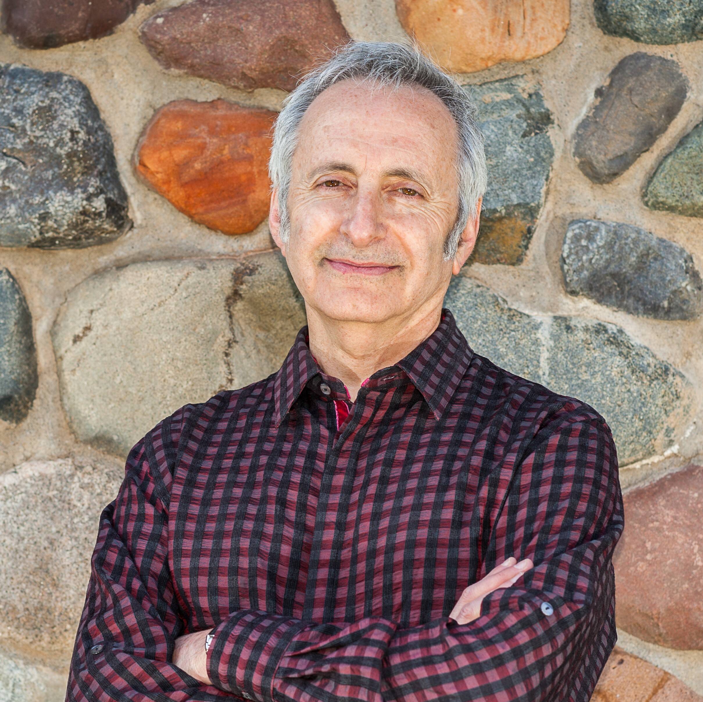 A man with gray hair stands with his arms crossed, smiling at the camera. He is wearing a maroon and black checkered shirt. The background features a rustic stone wall with various sized rocks in different colors, including brown, gray, and orange.