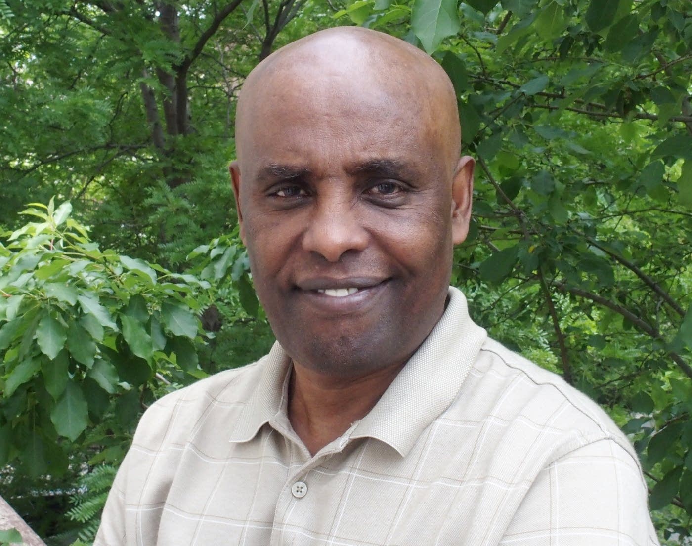 A man in a light-colored polo shirt smiles at the camera. He's standing outdoors with lush green foliage in the background.