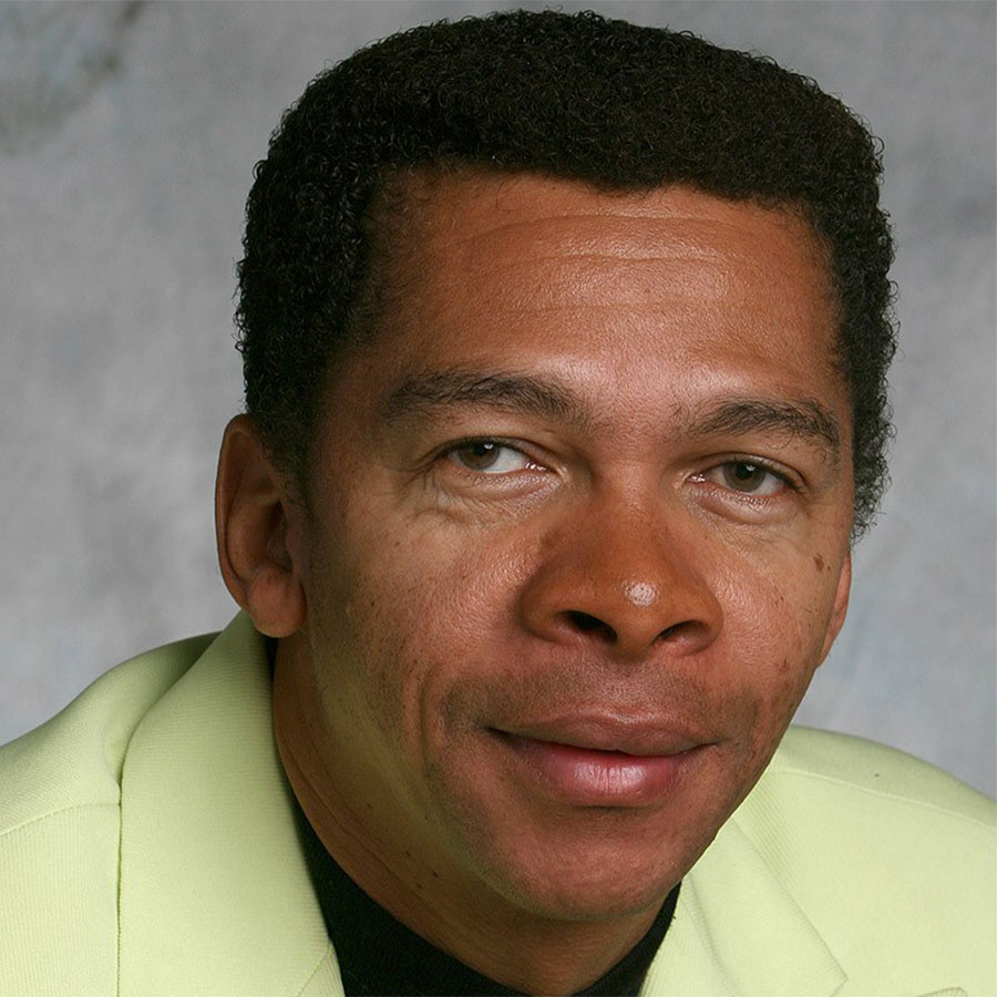 A man with short curly hair is wearing a pale green jacket and a black shirt. He has a neutral expression and is looking directly at the camera. The background is a soft, light-colored gray.