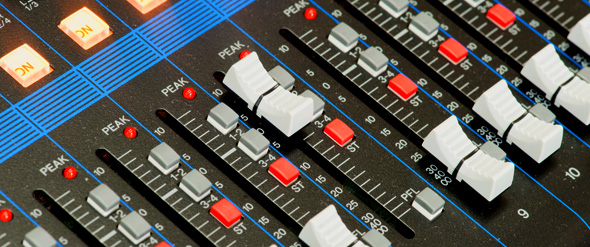 Close-up view of an audio mixing console with various sliders, buttons, and peak indicators. The controls include gray sliders and red buttons arranged in rows, surrounded by blue grid lines and LED indicators.