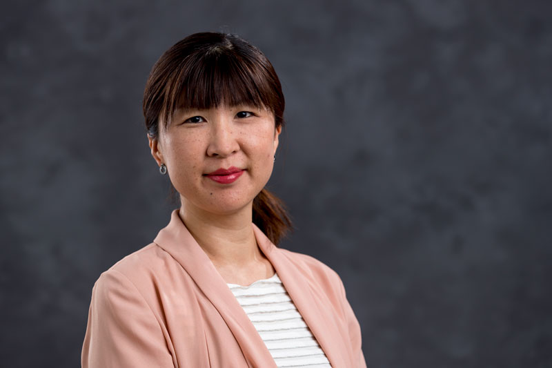 A woman with straight, dark hair tied back stands in front of a dark, blurred background. She is wearing a pink blazer over a white top and has a neutral expression.