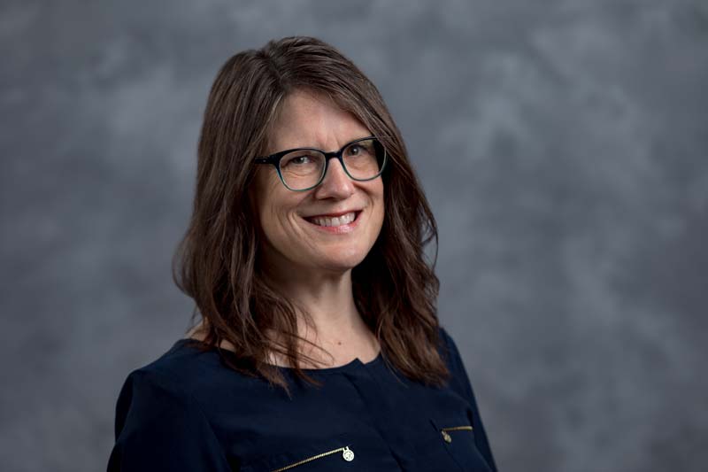 A person with long brown hair, wearing glasses and a dark blue blouse, is smiling against a gray, textured background.