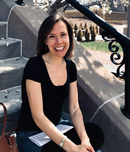 A person with shoulder-length light brown hair wearing a black short-sleeved shirt and jeans is sitting on outdoor steps, smiling at the camera. A handbag and some papers are beside them. It's a sunny day with trees and greenery in the background.