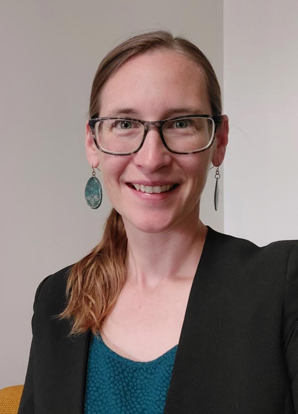 A person with long, light brown hair tied back, wearing glasses, drop earrings, and a dark blazer over a teal top, smiles at the camera. They are indoors against a plain light-colored background.