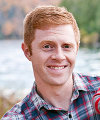 A smiling individual with short red hair wears a plaid shirt. The background is blurry, featuring greenery and a body of water.