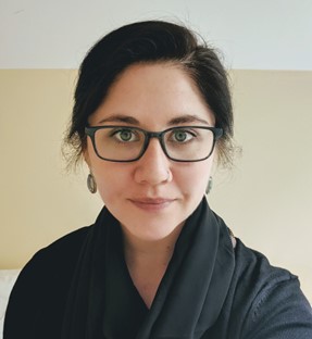 A person with dark hair, wearing glasses and a black scarf over a black top, looks directly at the camera. The background is a plain, light-colored wall.