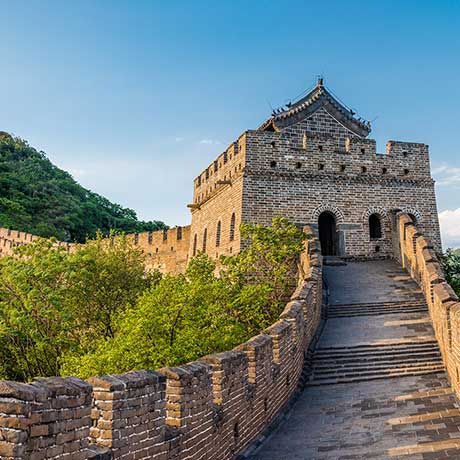 Image of the Great Wall of China, featuring a stone-walled section with a watchtower. The wall curves and rises along a lush, green hillside under a clear blue sky.