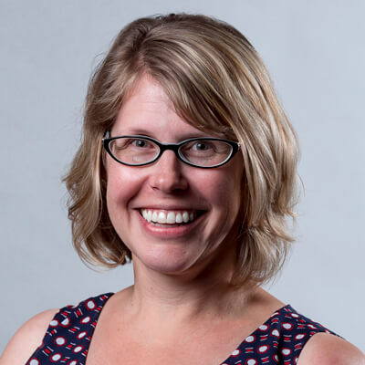 A woman with blonde hair and black-framed glasses is smiling at the camera. She is wearing a sleeveless top with a pattern of red and white circular shapes on a dark blue background. The background of the image is plain and light-colored.