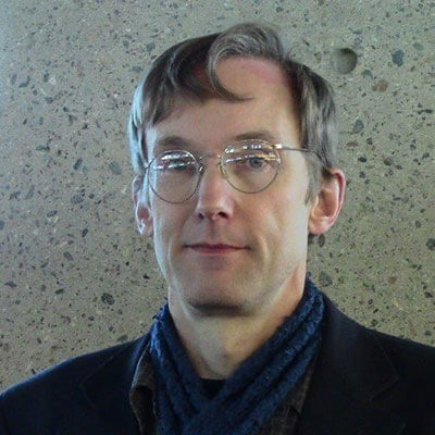 A person with light brown hair, wearing glasses and a blue scarf, is standing in front of a textured stone wall. He is facing the camera and wearing a dark suit jacket over a darker shirt.