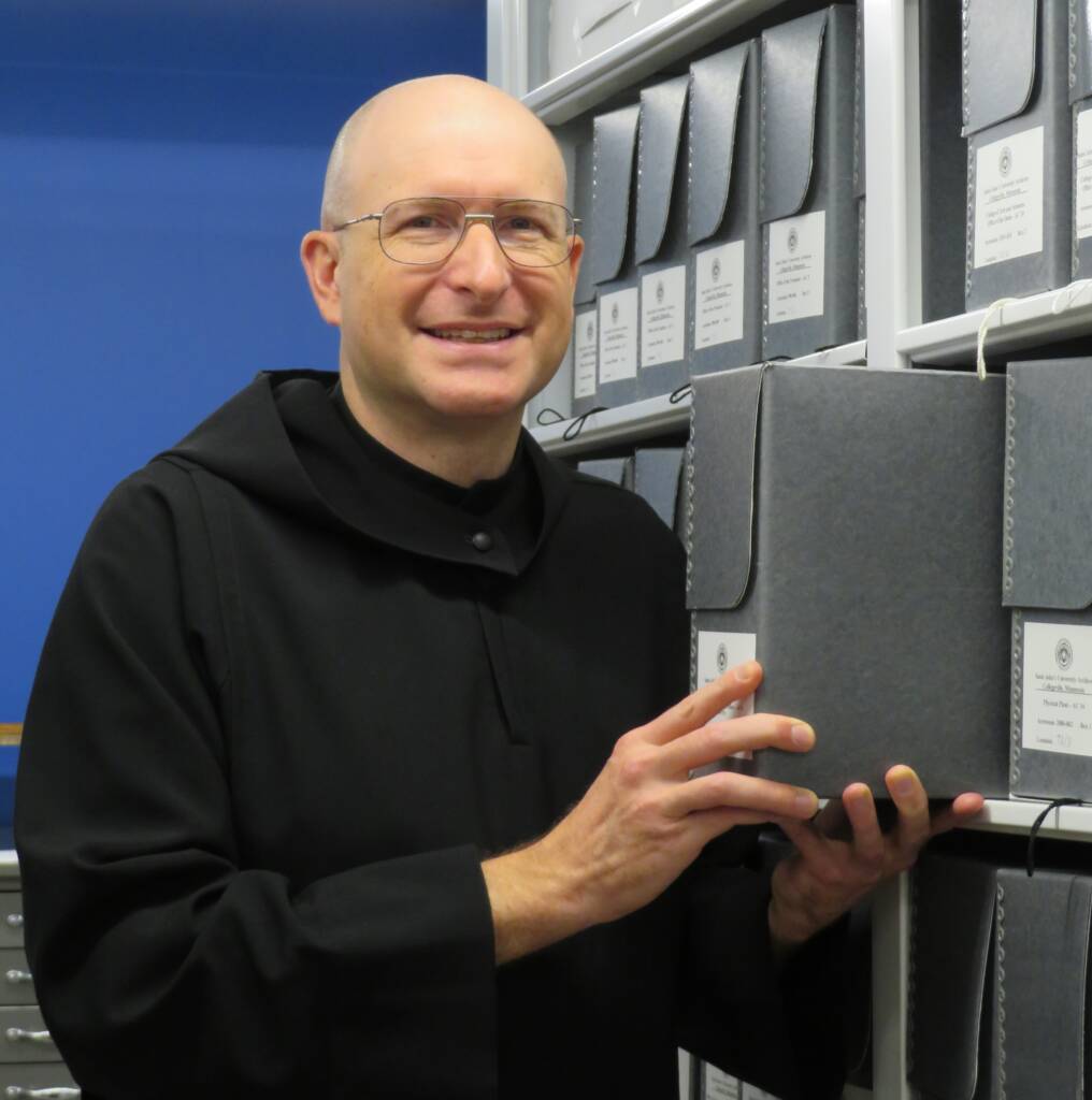 A person in a black robe, wearing glasses, stands in front of shelves filled with gray archival boxes. The person has a bald head and is smiling at the camera, holding one of the boxes. The background is a blue wall.