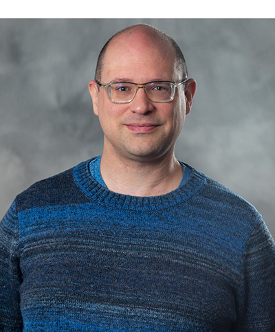 A person wearing glasses and a blue sweater stands against a gray, softly textured background, smiling slightly.