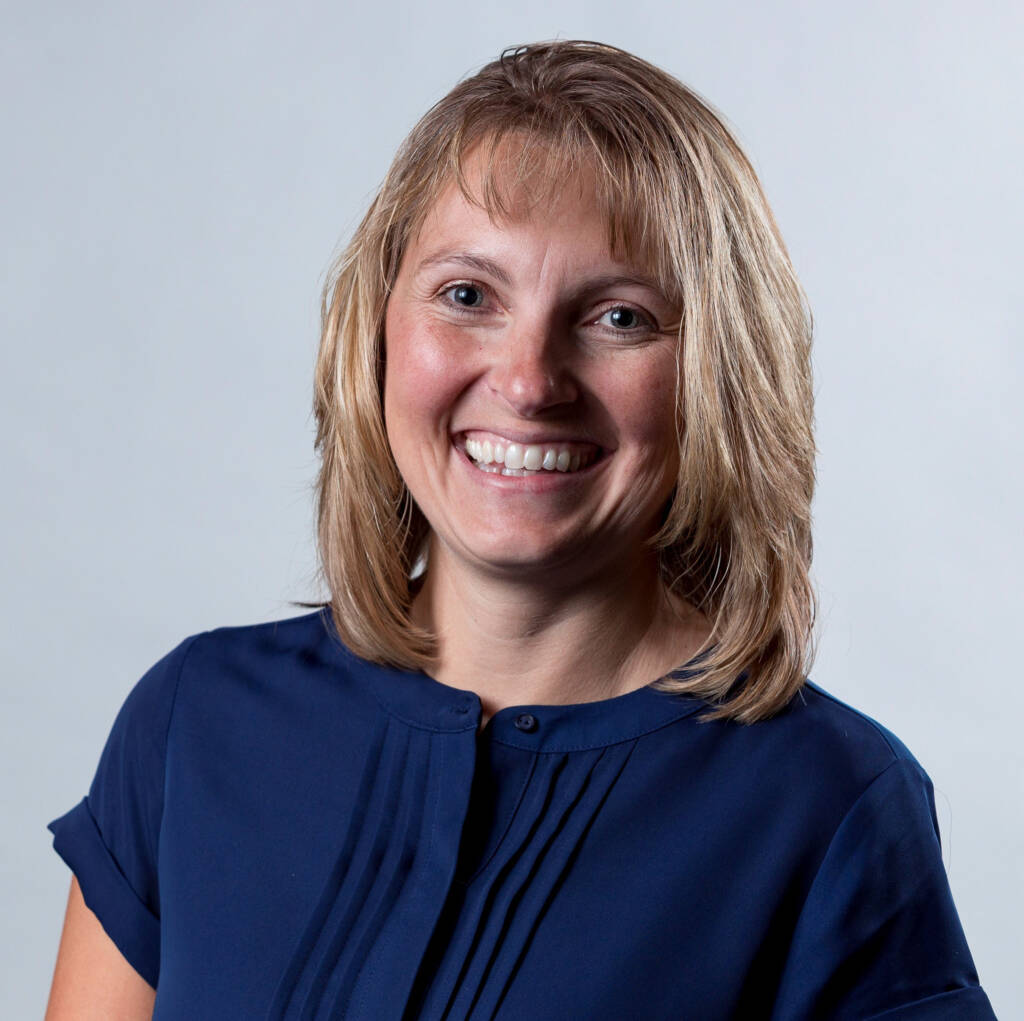 A woman with shoulder-length blonde hair is smiling at the camera. She is wearing a navy blue, short-sleeved top with subtle pleats on the front. The background is a plain light gray.