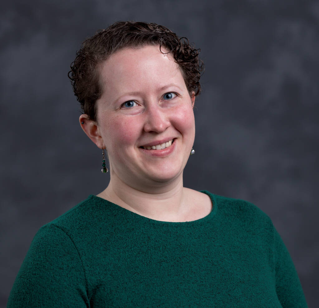 A person with short curly hair is smiling at the camera. They are wearing a green top and dangling earrings. The background is a neutral, dark gray color.