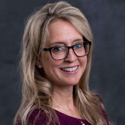 A woman with long blonde hair and glasses smiles at the camera. She is wearing a dark purple top against a dark gray background.