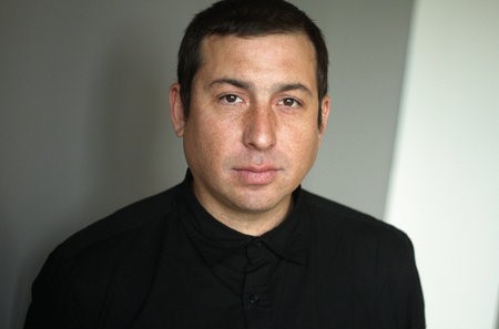 A person with short dark hair is wearing a black buttoned-up shirt and looking directly at the camera. The background is a neutral gray.