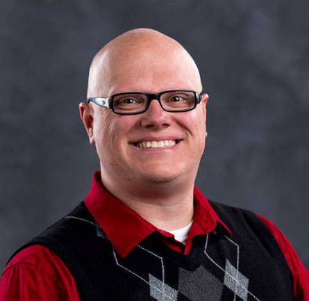 A bald individual wearing glasses smiles at the camera. They are dressed in a red shirt, a black sweater vest with a diamond pattern in gray and white. The background is a dark, neutral tone.