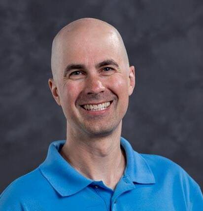 A bald man with a broad smile, wearing a light blue polo shirt, stands against a dark, blurred background.