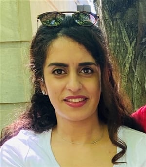 A woman with long, curly brown hair is smiling at the camera. She is wearing a white top and sunglasses resting on her head. She is standing outdoors, with a large tree and a building in the background.