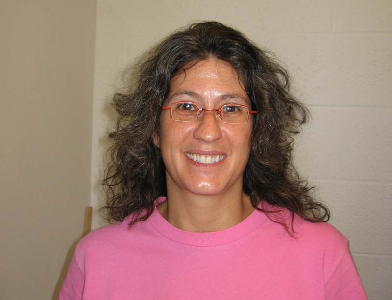 A woman with wavy, shoulder-length dark hair and red-framed glasses is smiling at the camera. She is wearing a pink shirt and standing against a plain, light-colored wall with a slightly textured surface.