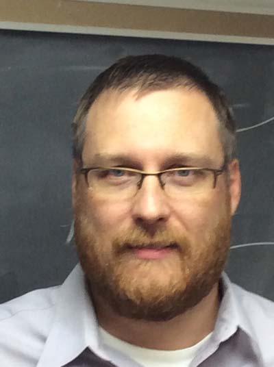 A man with short brown hair, a full beard, and glasses is pictured in front of a chalkboard. He is wearing a light-colored collared shirt and looking directly at the camera with a neutral expression.