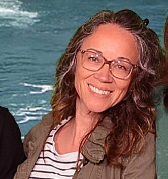 A smiling woman wearing glasses, a green jacket, and a striped shirt stands in front of a blue water background. She has wavy brown hair and is slightly angled toward the camera.
