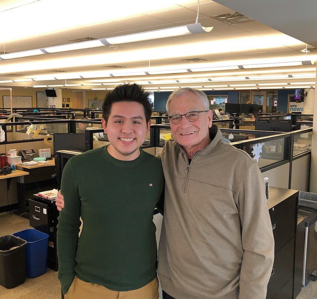 Two people are standing and smiling in a well-lit office. The younger person on the left is wearing a green sweater and tan pants, while the older person on the right is wearing a grayish zip-up sweater. Both have their arms around each other’s shoulders. Office cubicles and equipment can be seen in the background.