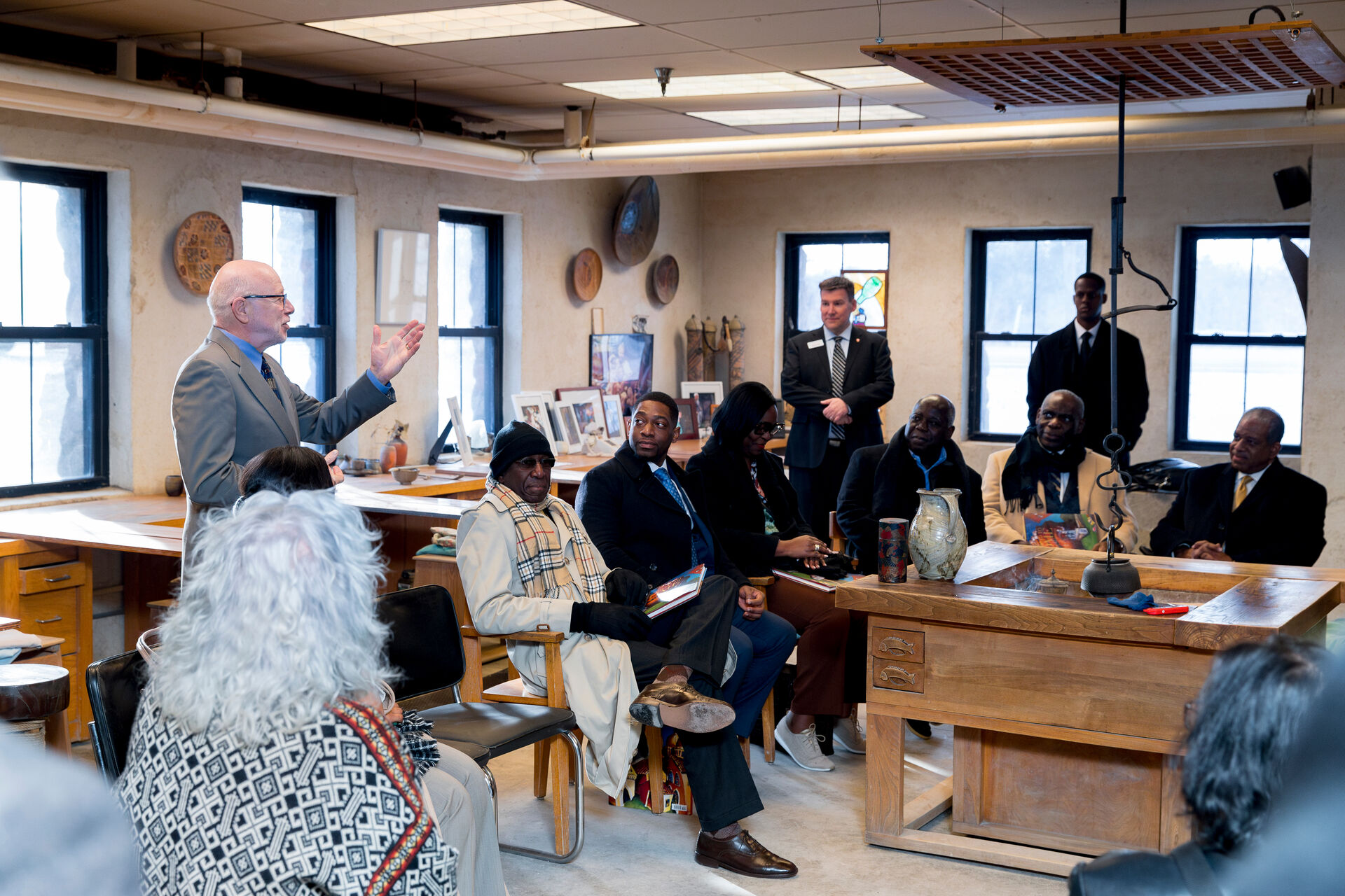 A group of people are gathered in a room with large windows and various artworks. An older man with glasses, standing and speaking, captures the group's attention. Some are seated while others stand around a large wooden table set with art supplies.