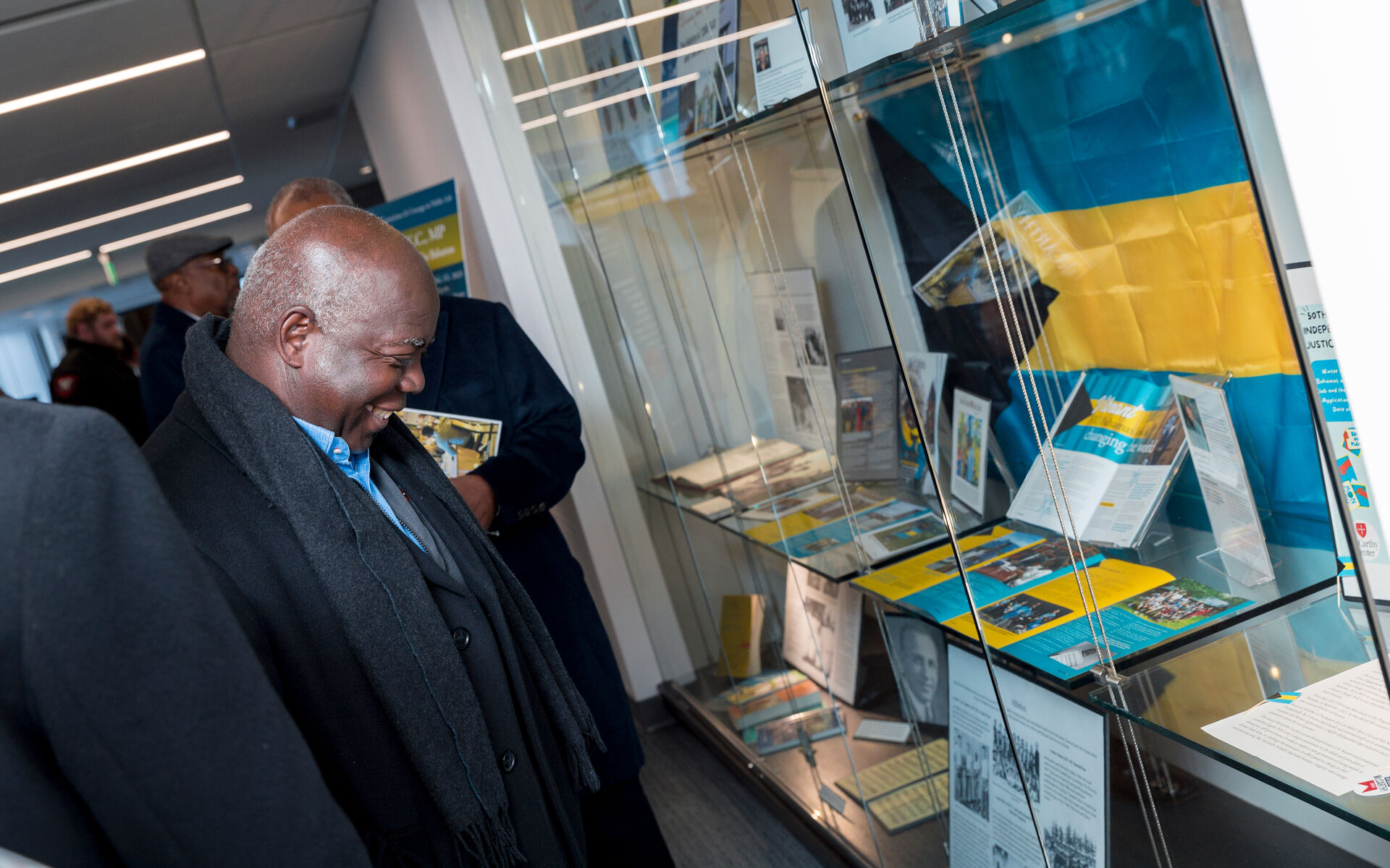 A man in a dark coat and scarf smiles while looking at a glass display case filled with documents and memorabilia. There is a large flag in blue and yellow colors partially visible in the background. Several other individuals are in the background observing the display.