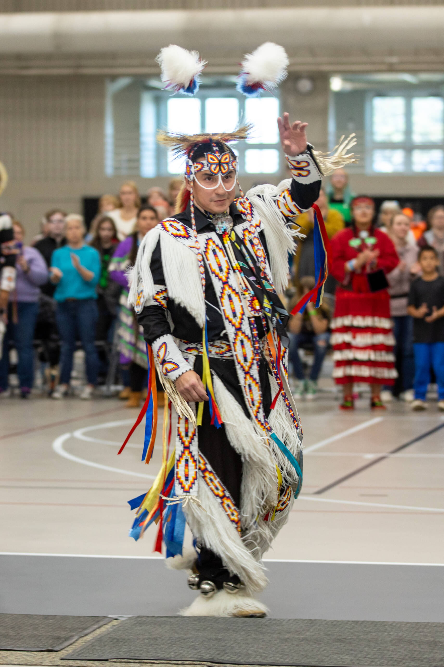 A person dressed in traditional regalia with colorful patterns and fringed elements performs in a gymnasium. They wear a headdress with white plumes and have beaded designs. A crowd watches in the background.