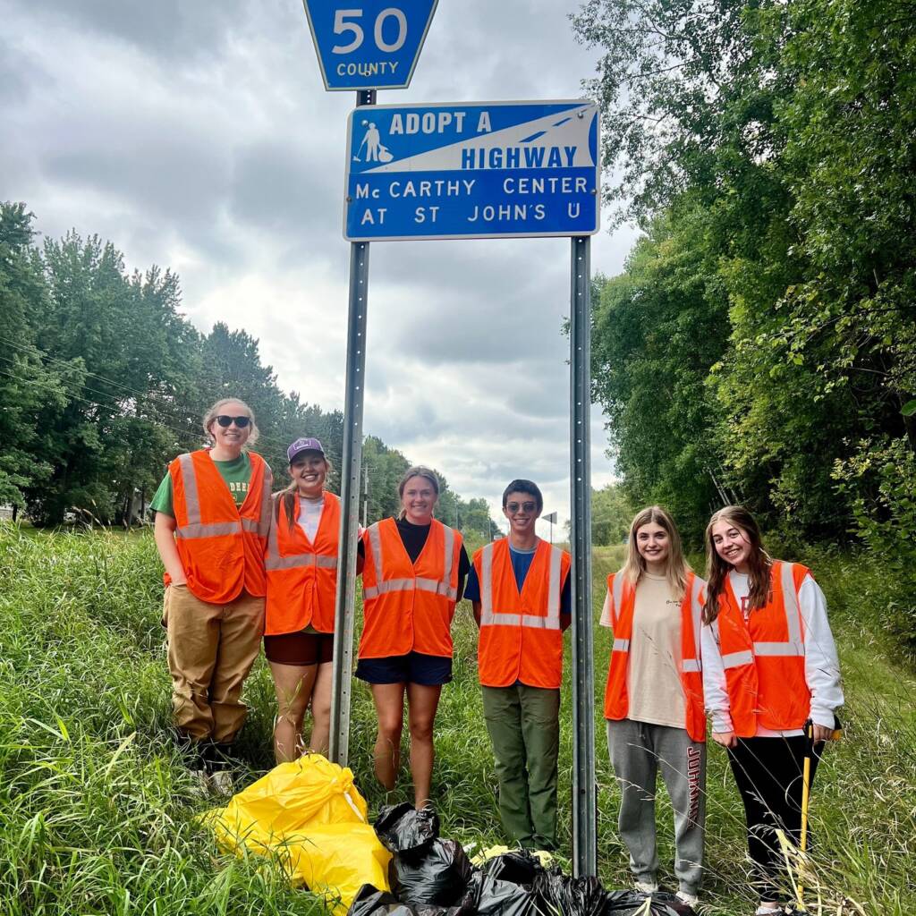 A group of six people wearing orange safety vests stand next to an "Adopt A Highway" sign on a grassy roadside. Several black trash bags are at their feet. Trees line the road, and the sky is cloudy.