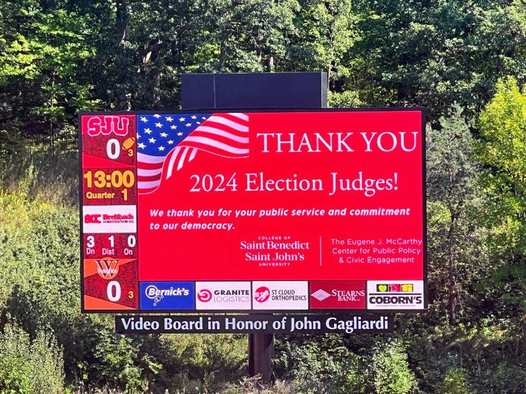 A red digital billboard in a green, wooded area reads, "Thank you 2024 Election Judges!" with an American flag design. It includes score graphics and school logos, and recognizes public service and commitment to democracy.