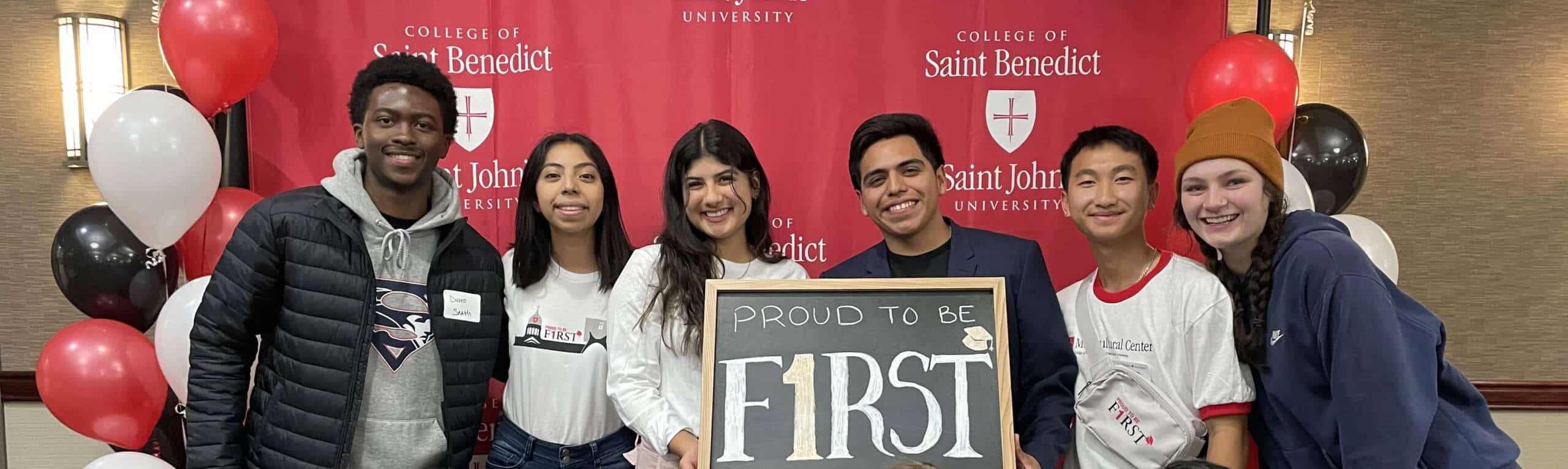 A group of six diverse students stands smiling in front of a red backdrop with "College of Saint Benedict" and "Saint John" logos. They hold a chalkboard sign reading "PROUD TO BE FIRST." The students are surrounded by red, white, and black balloons.