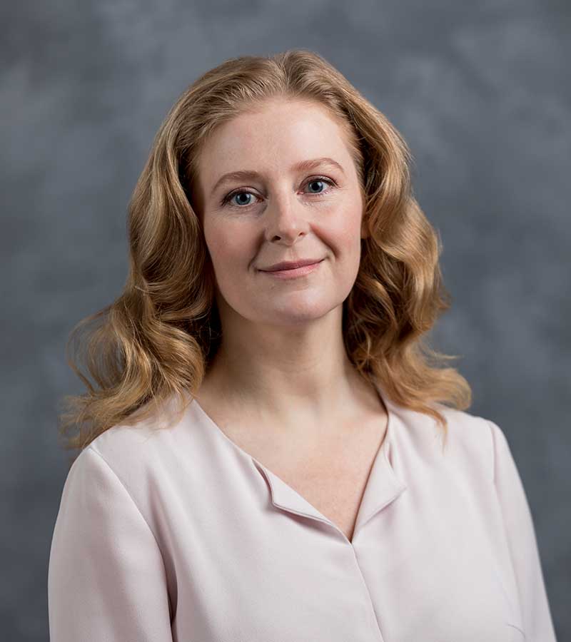 A woman with shoulder-length, wavy blonde hair is smiling slightly. She is wearing a light pink blouse and is posed against a grey background.