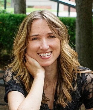 A person with long, wavy hair and a black top is smiling warmly at the camera. They are sitting outdoors with one hand resting on their cheek and green foliage in the background.