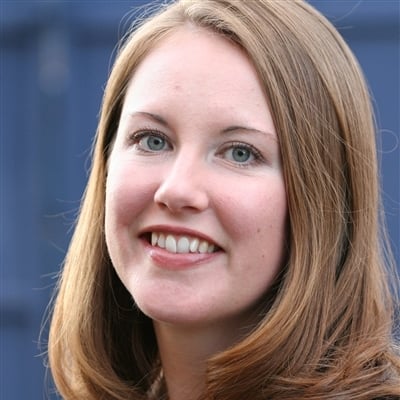 A woman with light skin and shoulder-length light brown hair is smiling. She is wearing a light-colored top. The background is slightly blurred with a deep blue hue.