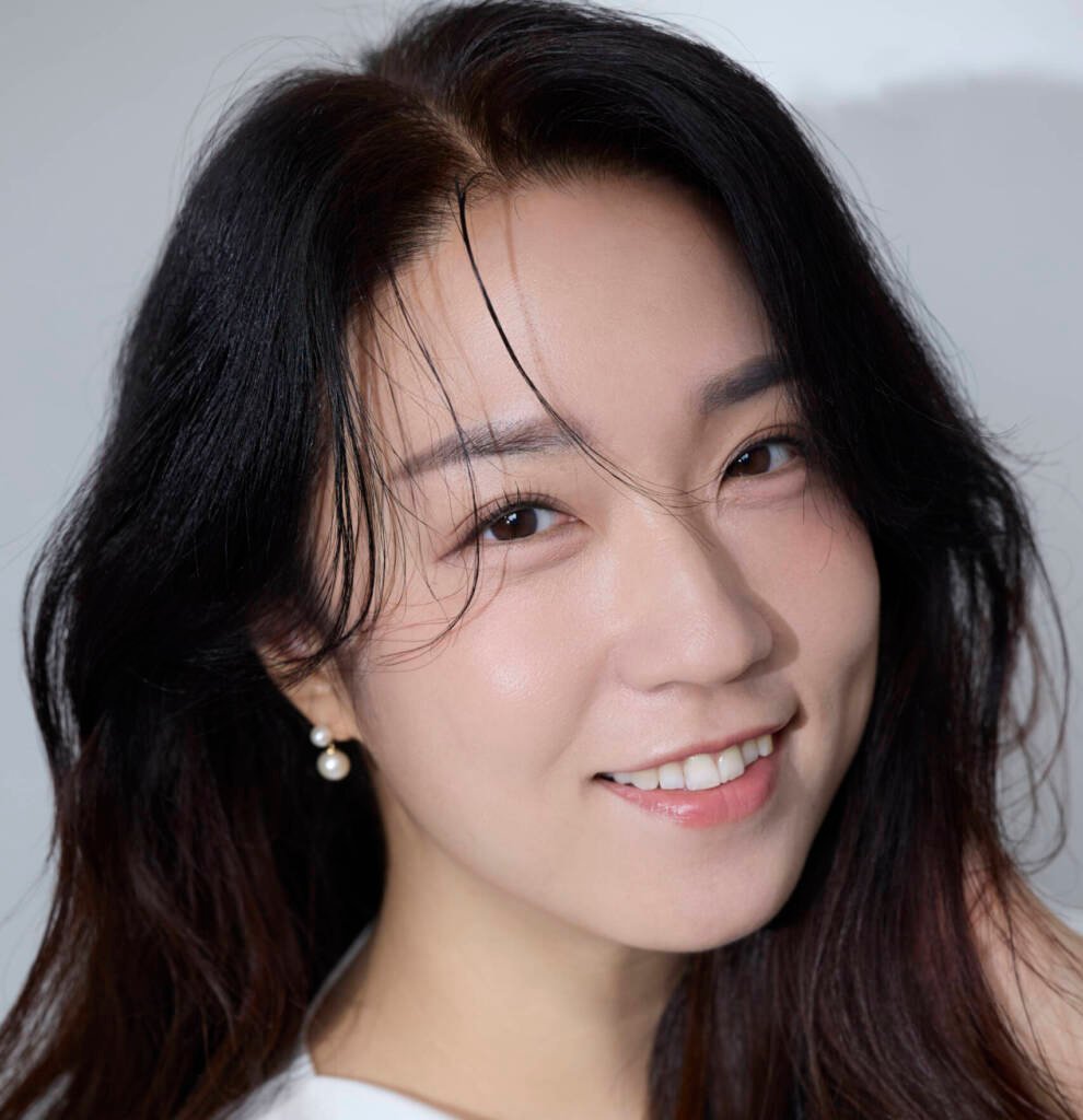 A woman with long, dark hair smiles at the camera. She is wearing a white top and pearl earrings. Her hair is slightly tousled, framing her face gently. The background is plain and light-colored, emphasizing her bright and cheerful expression.