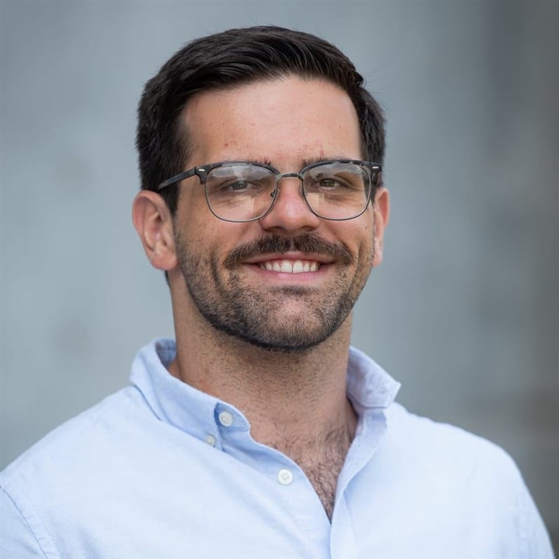 A man with brown hair, a beard, and glasses is smiling at the camera. He is wearing a light blue button-up shirt and is standing in front of a neutral, blurred background.