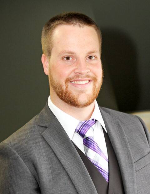 A man with short hair and a trimmed beard smiles at the camera. He is wearing a gray suit, a white dress shirt, and a purple striped tie, with a dark vest underneath. The background is a dark, solid color.