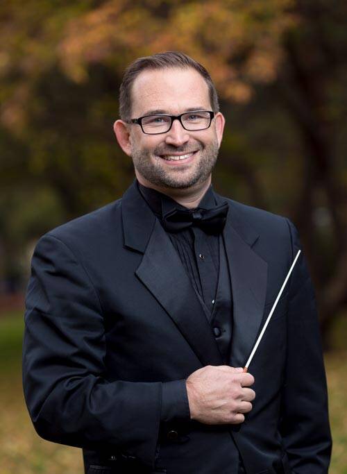 A man wearing a black suit and bowtie smiles while holding a conducting baton. He has short hair, glasses, and a neatly trimmed beard. The background is outdoors with blurred trees and fallen leaves, suggesting an autumn setting.